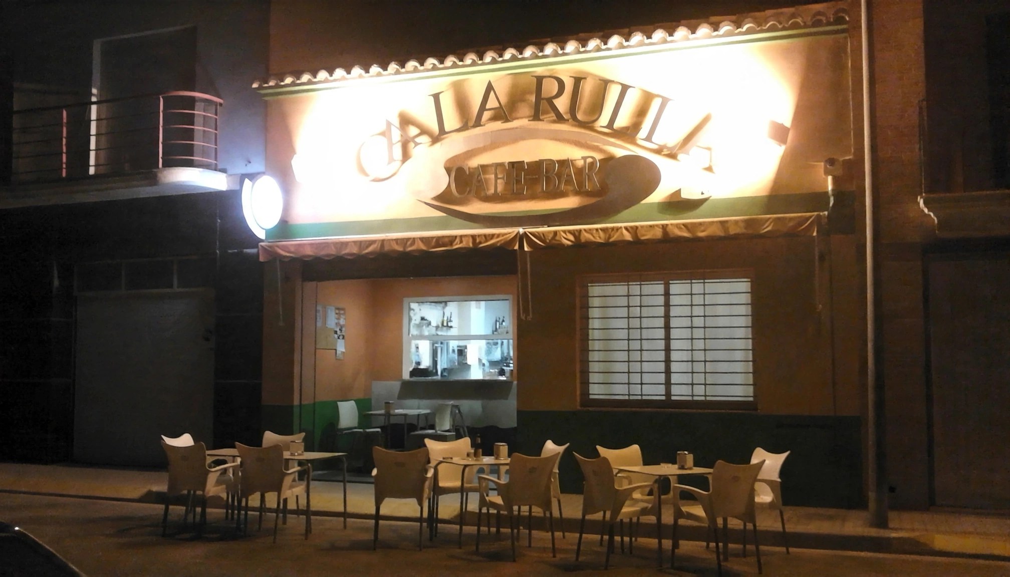 Fachada iluminada de Ca la Rulla Café-Bar por la noche, con su terraza y mesas preparadas en Bellreguard